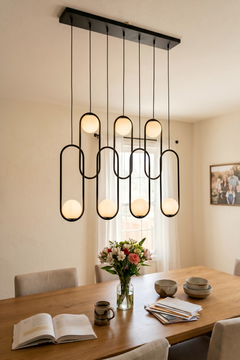 Modern minimalist chandelier illuminating a wooden dining table set with flowers and an open cookbook in a sunlit dining room